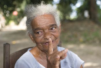 Senhora negra e idosa sentada numa cadeira num quintal contando uma história