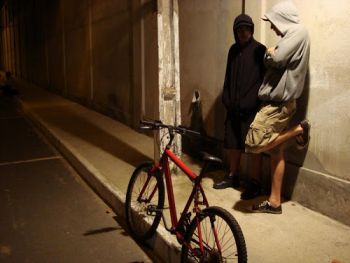 Dois jovens encostados em uma parede conversando numa calçada com uma bicicleta em frente